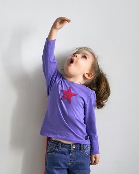 A young child in a purple star shirt, jeans, and a red cape reaching upward against a light wall