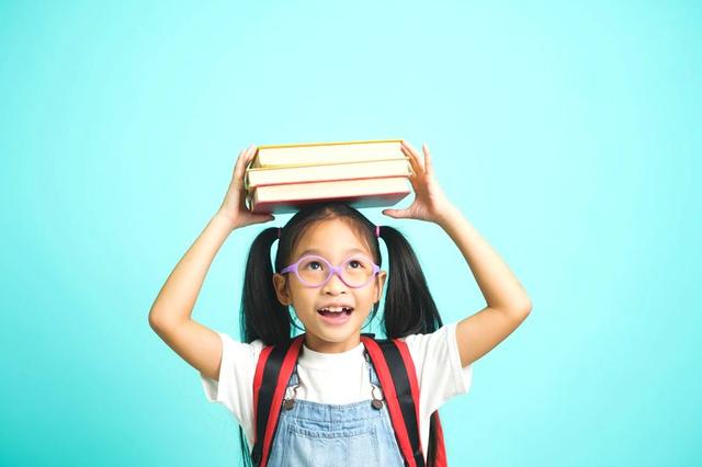 A child balancing books on their head, wearing purple glasses and a red backpack